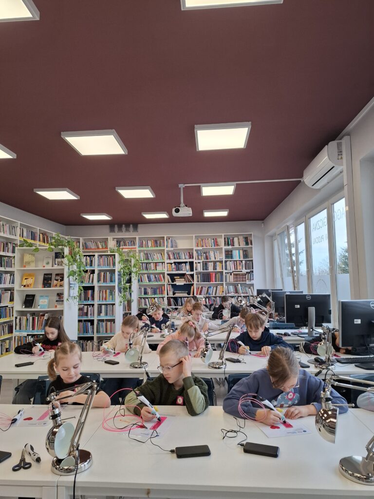 Grupa dzieci w bibliotece podnosi do góry zakładki do książki wykonane długopisami 3D.