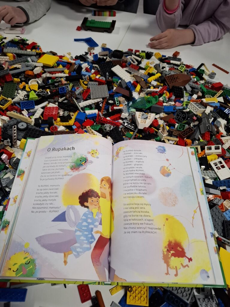 Zdjęcie przedstawia otwartą kolorową książkę dla dzieci, leżącą na stole, wokół której rozsypane są liczne klocki LEGO w różnych kolorach i kształtach. Na ilustracji w książce widzimy uśmiechniętą kobietę i dziecko w niebieskiej piżamie w chmurki, trzymające poduszkę. Obok nich unoszą się kolorowe, puszyste stworki. Tekst w książce jest w języku polskim i zatytułowany "O Rupakach". Dookoła książki widać dzieci bawiące się klockami LEGO — widać ich ręce i fragmenty ubrań.