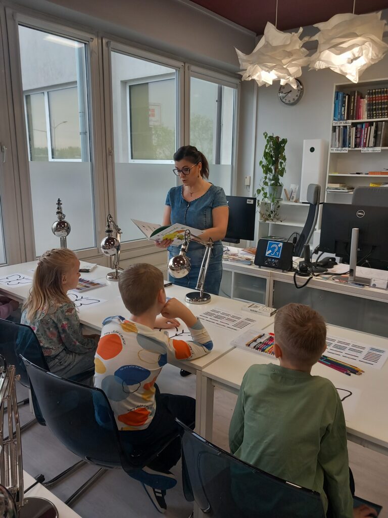 Na zdjęciu widoczna jest kobieta czytająca dzieciom książkę w nowocześnie urządzonej sali bibliotecznej. Kobieta stoi przy biurku, trzyma otwartą książkę i zwraca się do trójki dzieci siedzących przy stołach. Uczniowie uważnie słuchają, przed nimi leżą kartki z konturami motyli oraz zestawy kredek. Na stołach znajdują się także małe lampki oraz karty z kolorowymi kodami. W tle widać regały z książkami, monitory komputerów i duże okna wpuszczające naturalne światło. Atmosfera na zdjęciu jest spokojna i sprzyjająca nauce.