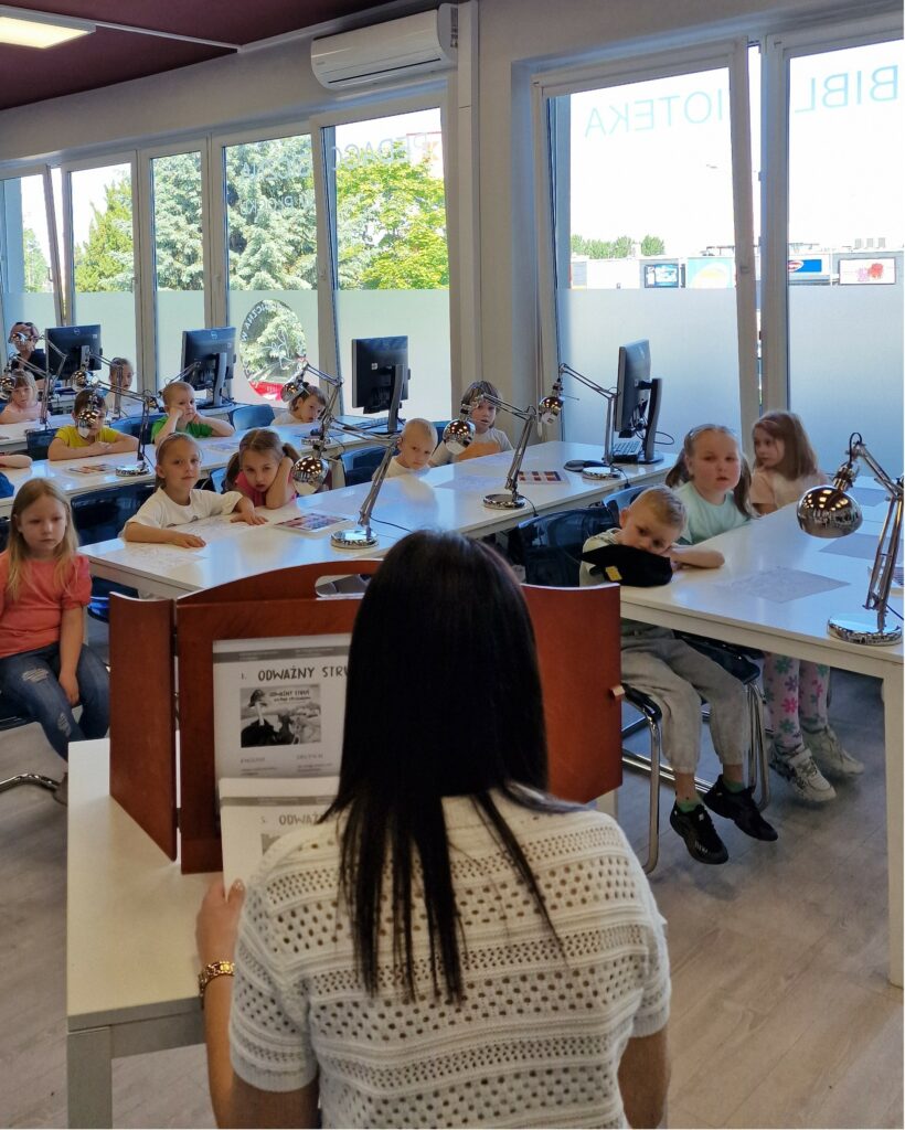 Dzieci z przedszkola słuchają bajki „Odważny struś zostaje strażakiem” prezentowanej w teatrzyku kamishibai w bibliotece.