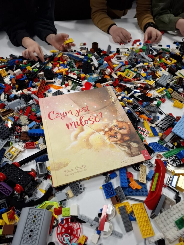 Na stole leży książka pt. „Czym jest miłość?” otoczona dużą ilością klocków LEGO, wokół których widać dziecięce ręce.
