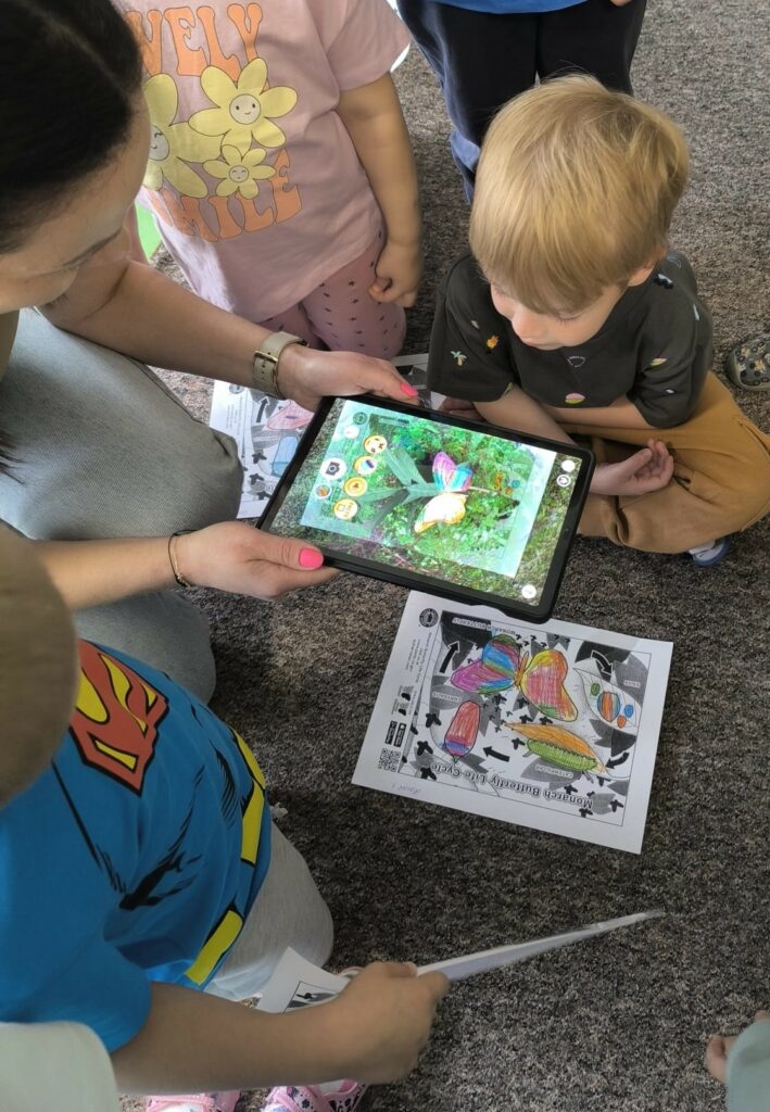 Na zdjęciu widać dzieci, które podczas zajęć oglądają na tablecie animację kolorowego motyla, który ożył dzięki aplikacji Quiver.