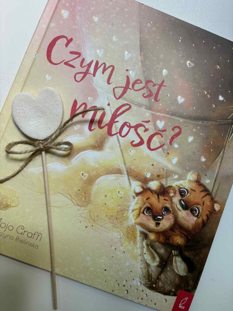 Na zdjęciu znajduję się okładka książki Czym jest miłość? Mojo Graffi z ilustracją dwóch uśmiechniętych tygrysków siedzących razem w koszu balonu.