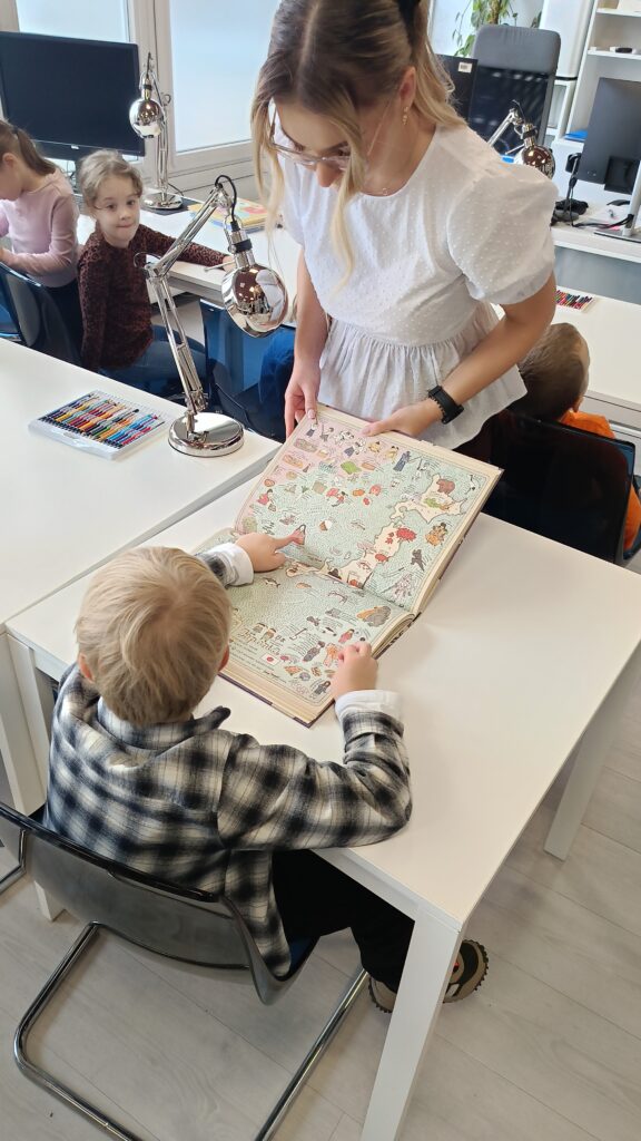 Na zdjęciu widać kobietę pokazującą dziecku otwartą książkę z kolorową mapą. Chłopiec siedzi przy białym stoliku i wskazuje palcem na fragment ilustracji. W tle widać inne dzieci zajęte pracą przy stolikach.