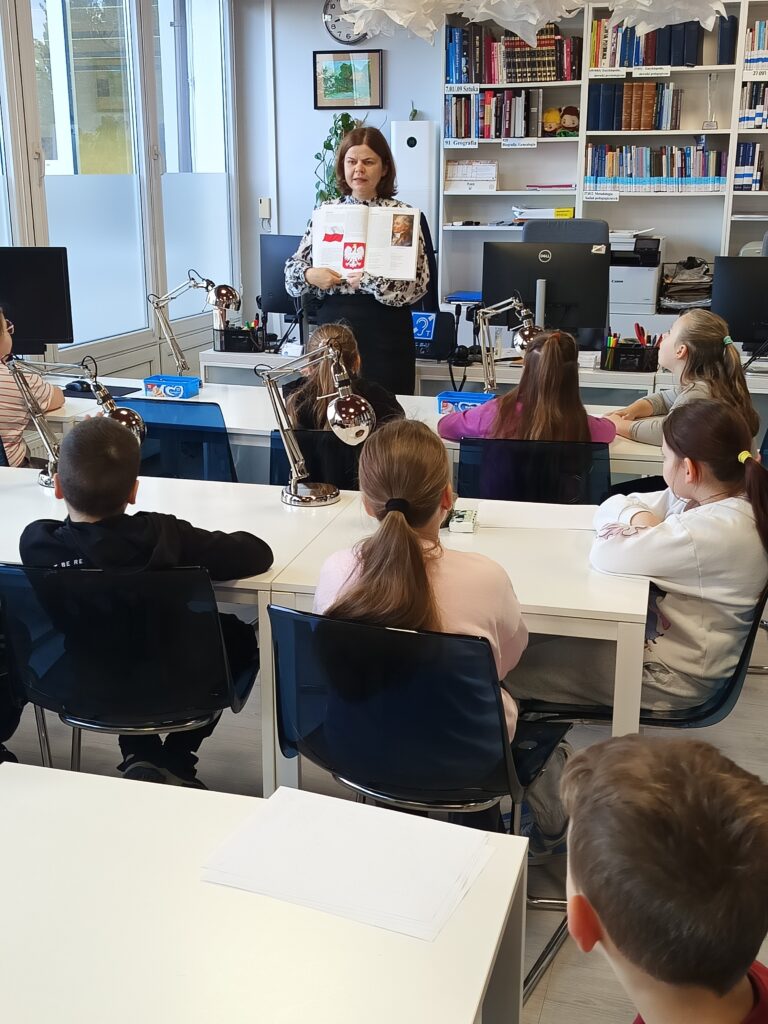 Nauczycielka stoi przed uczniami klasy 3 SP, którzy siedzą w ławkach. Kobieta trzyma w rękach otwartą książkę i pokazuje dzieciom ilustracje.