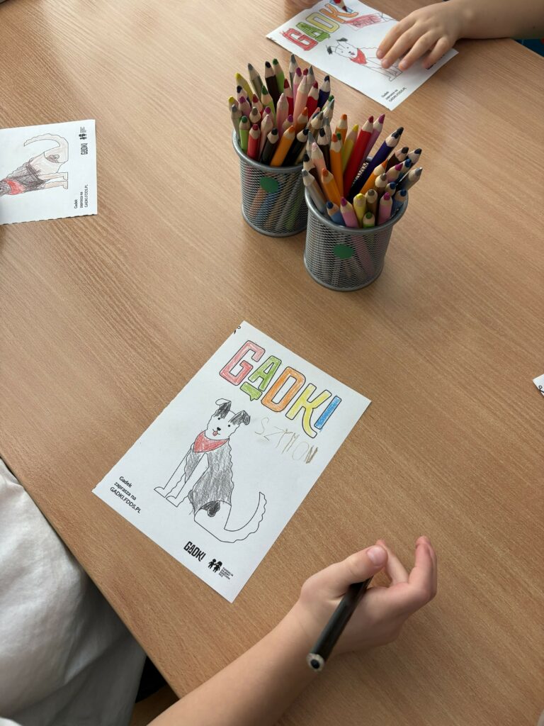 Na zdjęciu widać dzieci, które kolorują rysunki psa z napisem GADKI przy stole, na którym stoją pojemniki z kolorowymi kredkami.