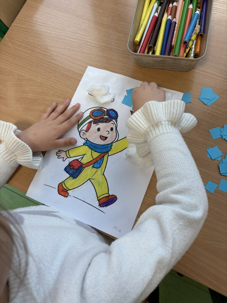 Na zdjęciu widać dziecko, które siedzi przy stoliku i ozdabia kolorowankę przedstawiającą uśmiechniętego chłopca w pilotce. 
