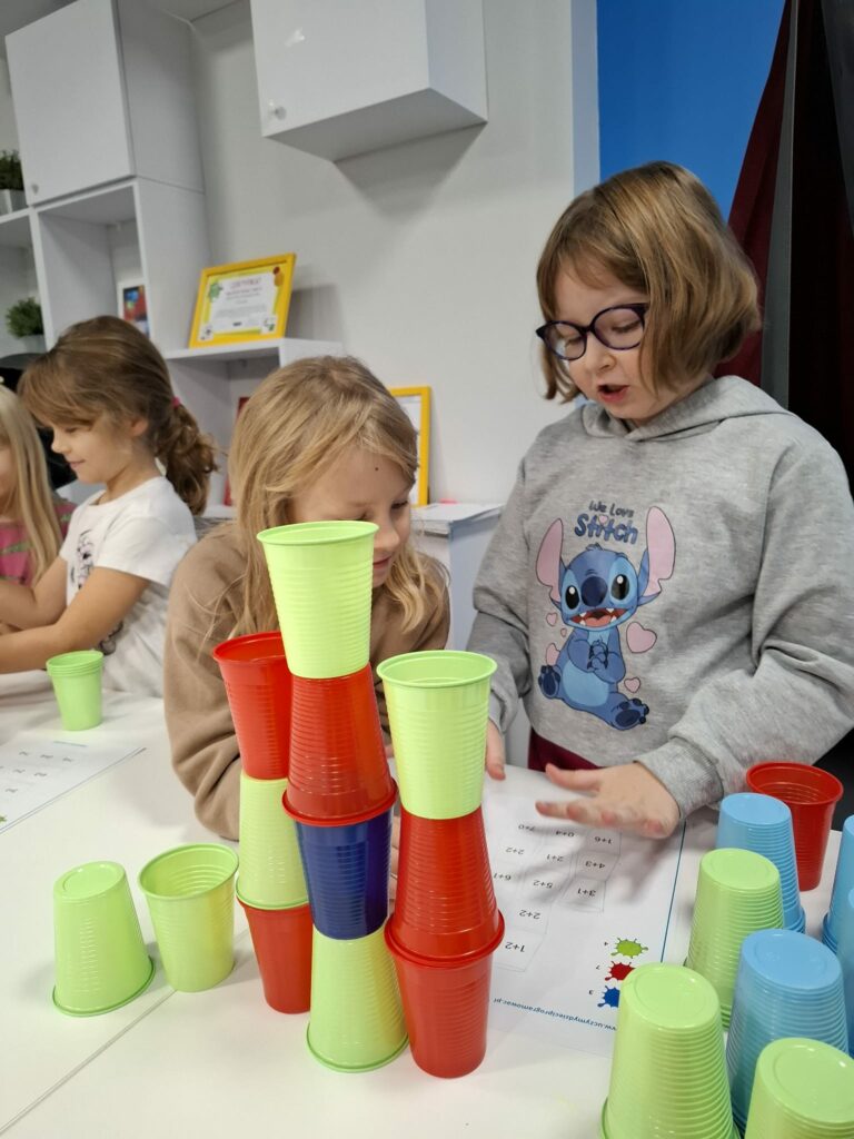 Na zdjęciu widać dzieci pracujące wspólnie przy stoliku, układające kolorowe plastikowe kubki według wzoru; obok nich leżą karty z zadaniami.
