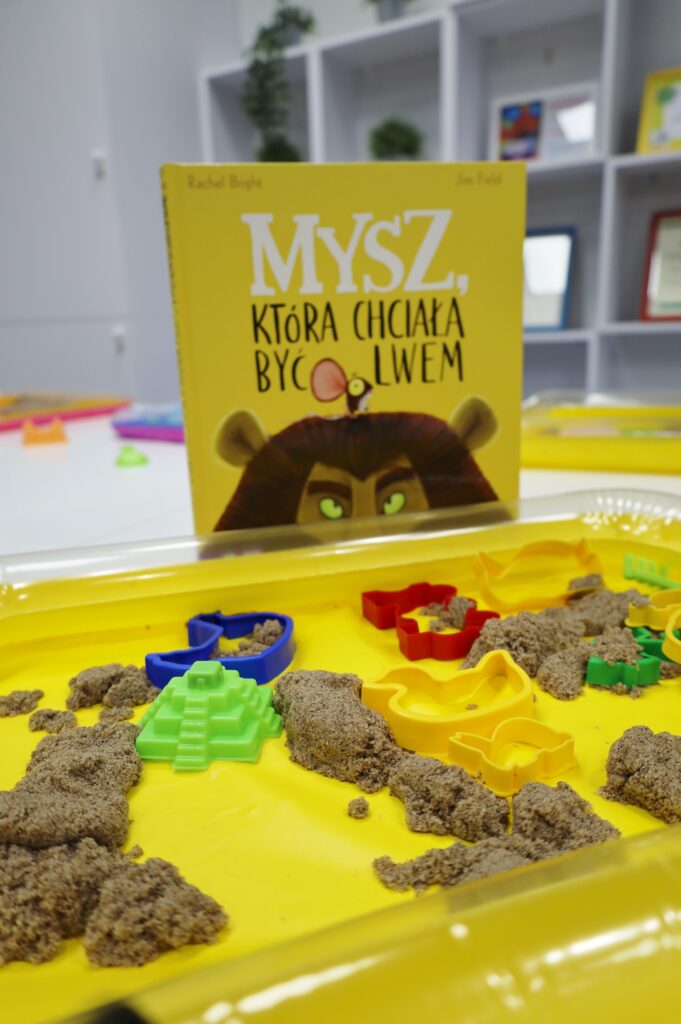 Na zdjęciu widać książkę „Mysz, która chciała być lwem” ustawioną na stole w bibliotece. Przed nią znajduje się żółta taca z kinetycznym piaskiem oraz kolorowymi foremkami w kształcie zwierząt i budowli, które służą dzieciom do lepienia figurek podczas zajęć czytelniczych. W tle widoczne są półki z książkami i dekoracjami