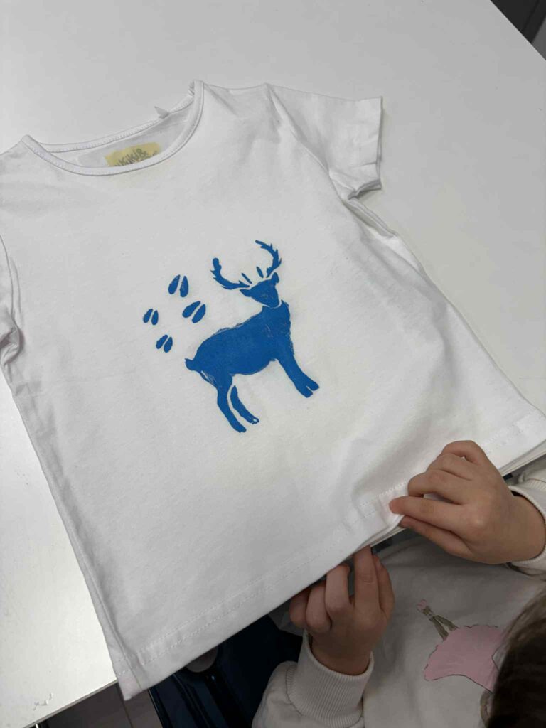 Biała dziecięca koszulka z niebieskim nadrukiem jelenia i śladów kopyt, trzymana w dziecięcych dłoniach.