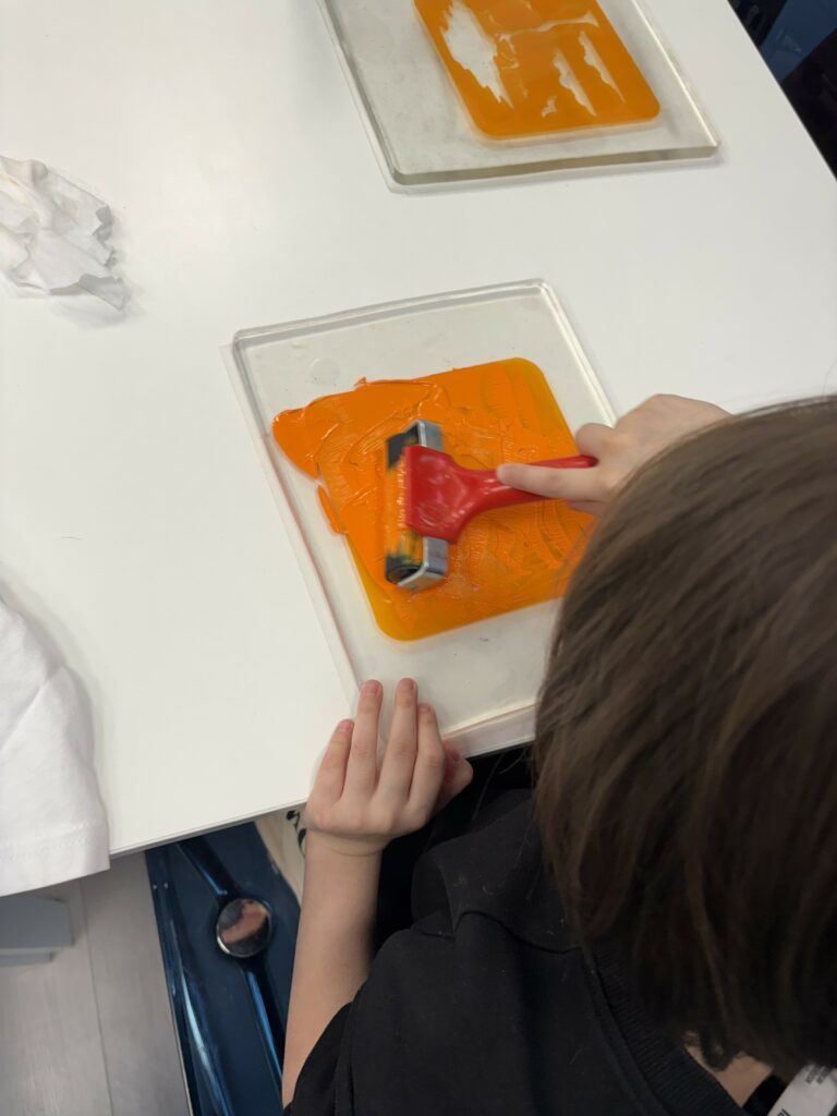 Na zdjęciu widać dziecko rozprowadzające pomarańczową farbę wałkiem na plastikowej płytce.