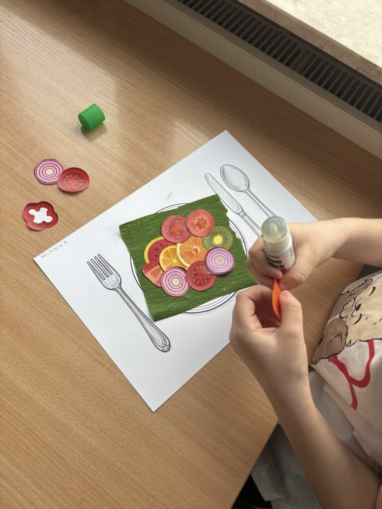 Na zdjęciu widać dziecko przyklejające papierowe warzywa i owoce na kartce z narysowanym talerzem i sztućcami. 