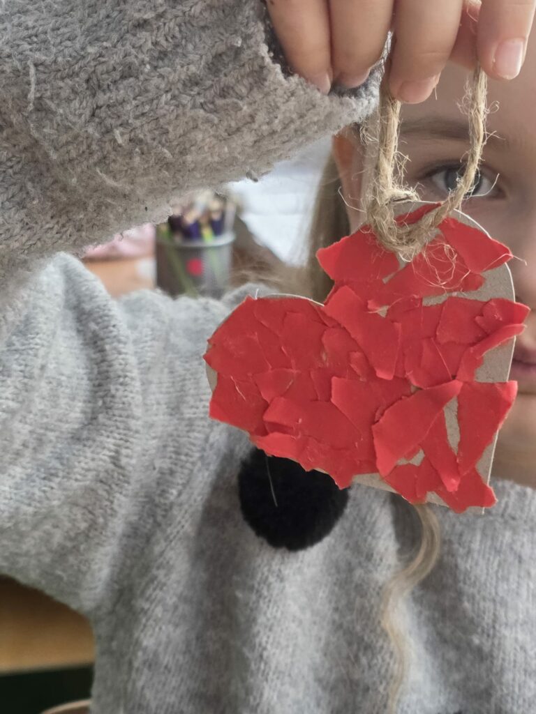 Dziecko trzyma w dłoni własnoręcznie wykonane serduszko wyklejone czerwonym papierem, zawieszone na sznurku.