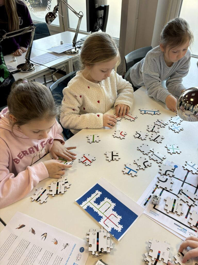 Na zdjęciu widać troje dzieci siedzących przy stole podczas zajęć edukacyjnych, które układają puzzle z kodami do programowania ozobotów.