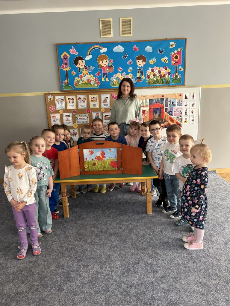 Na zdjęciu widać nauczycielkę stojącą z grupą dzieci w sali przedszkolnej przy stoliku z drewnianą skrzynią.