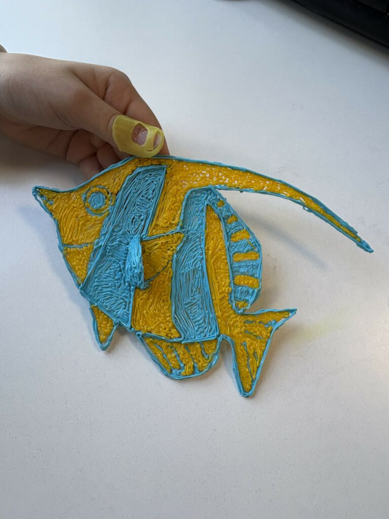 Na zdjęciu widać rękę dziecka trzymającą płaską figurkę ryby wykonaną z filamentu przy użyciu długopisu 3D.