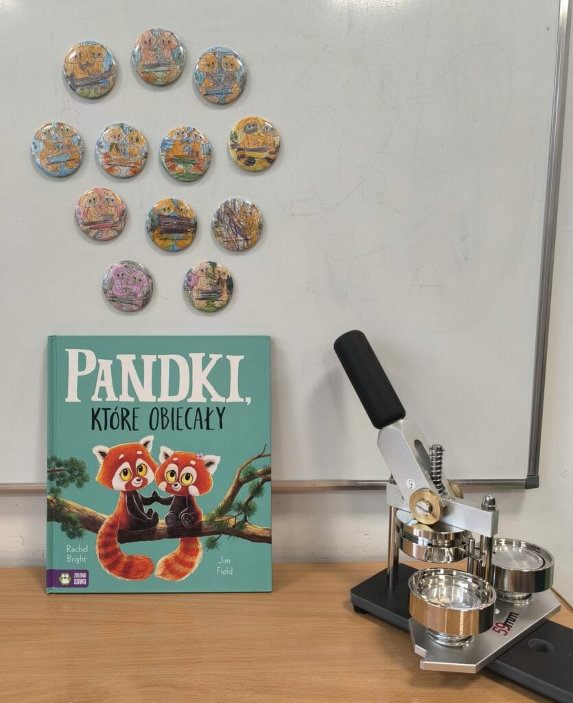 Na zdjęciu widać książkę „Pandki, które obiecały” autorstwa Rachel Bright i Jima Fielda stojącą na biurku. Obok znajduje się prasa do wykonywania magnesów, a nad książką na tablicy umieszczone są okrągłe magnesy z pokolorowanymi przez dzieci ilustracjami pandek.