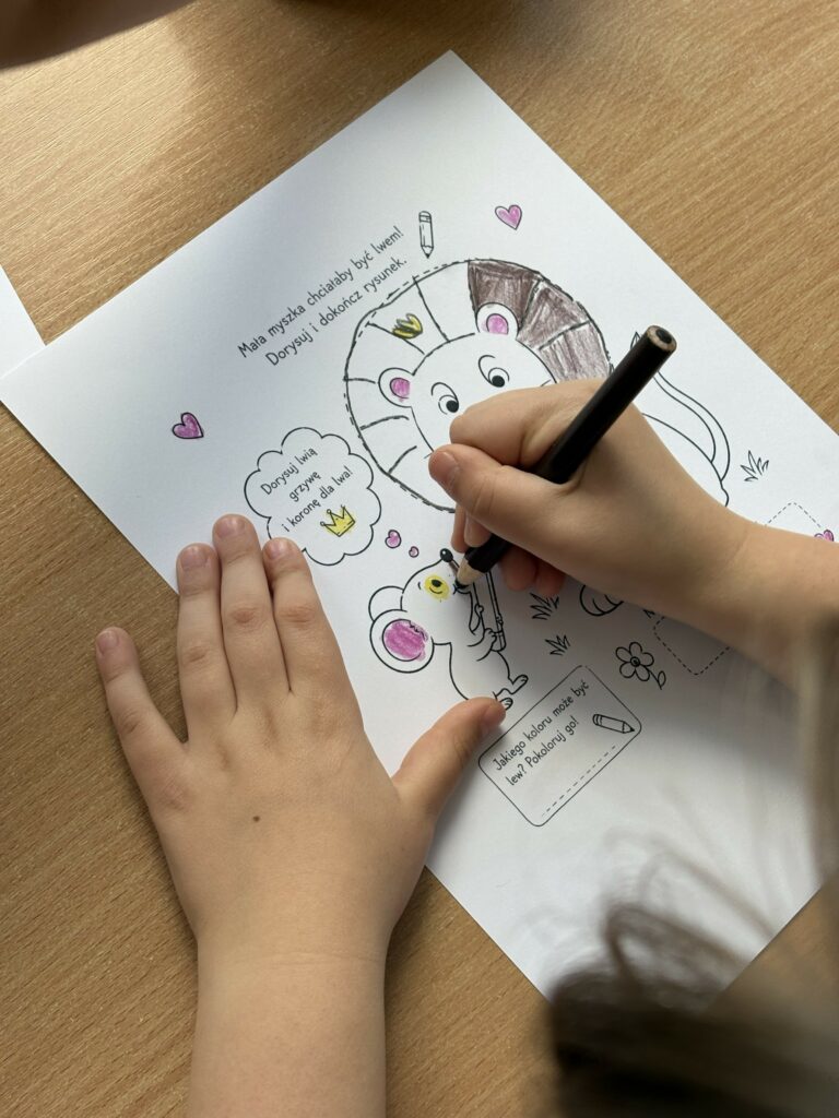 Na zdjęciu widać dziecko kolorujące kartę pracy inspirowaną książką „Mysz, która chciała być lwem”, na której dorysowuje lwu grzywę i koronę oraz ozdabia ilustrację kredkami.