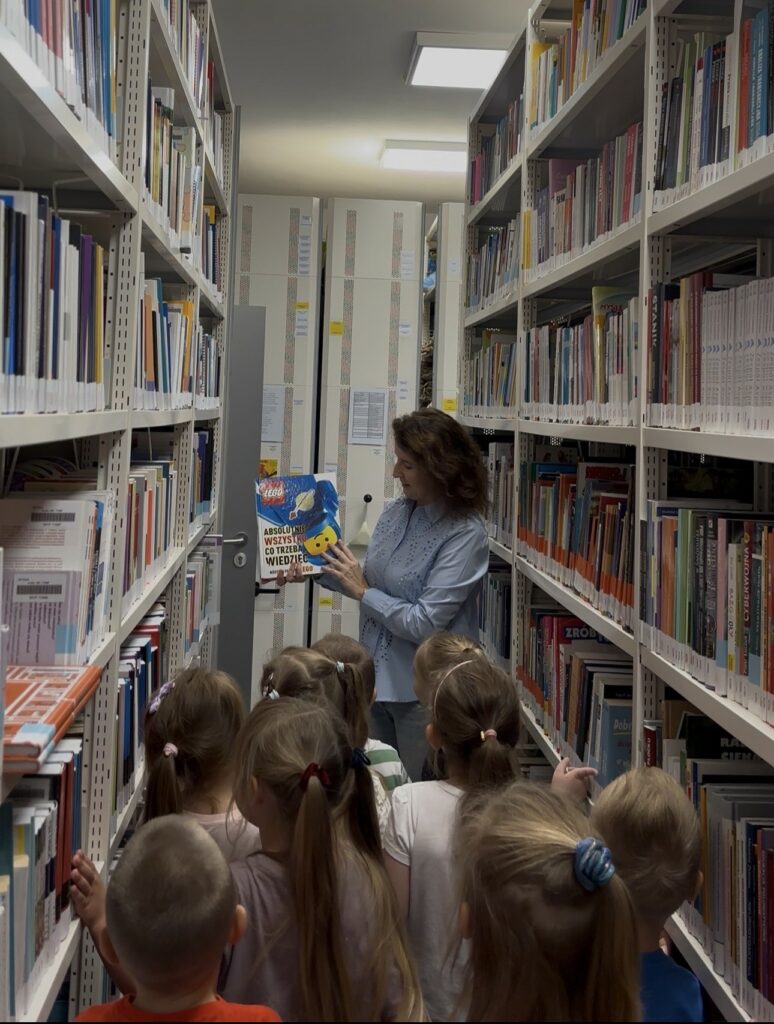 Na zdjęciu widać bibliotekarkę prezentującą książkę grupie dzieci wśród regałów z książkami w bibliotece.